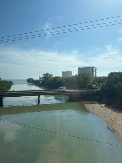 This image depicts a riverine and coastal industrial landscape within Kholodnyi Rodnik, Russia, observed during daylight hours under a clear blue sky with sparse, wispy white clouds. Three dark horizontal power lines traverse the upper portion of the sky.
A wide river, characterized by greenish-brown water reflecting the sky, occupies the middle ground and foreground. It flows from the viewer's right to left, emptying into a larger body of water, identified as the Black Sea, visible on the horizon. A concrete or metal bridge with pedestrian railings spans the river. A white van with a red stripe along its side is present on the bridge, traveling from right to left. A single pedestrian, a dark-clad individual, walks on the left side of the bridge, moving from left to right. Support pylons for the bridge are partially submerged in the river.
On the river's right bank in the foreground, a strip of coarse, light-colored gravel forms the shoreline. A rectangular, light-colored object, possibly a container or light fixture, rests on this gravel strip near the water's edge. Both river banks are densely covered with green trees and vegetation.
On the right bank, beyond the bridge and partially obscured by trees, stand two large, light-colored cylindrical industrial tanks or silos. The tank on the right is taller and features an external, spiraling metal staircase on its side. Industrial infrastructure and what appear to be older, multi-story structures are clustered near the river's mouth, where it meets the sea. These structures, some seemingly dilapidated, are built close to or directly over the water. A long, narrow structure, possibly a pier or breakwater, extends from the left bank into the sea. The sea itself appears lighter blue-green than the river.