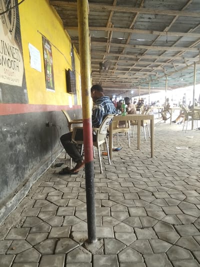 The image depicts an indoor or semi-outdoor casual dining or bar area, characterized by a relaxed atmosphere and simple construction. The overall setting suggests a public space, possibly a pub or local eatery.

In the foreground, a prominent yellow pole, painted with a black and red stripe at its base, stands vertically, partially obstructing the view. The ground is paved with interlocking grey concrete blocks forming a tessellated pattern. Along the left side of the frame runs a wall, painted bright yellow on the upper section and dark grey or black on the lower part, separated by a thin red horizontal stripe.

Mounted on the yellow wall are several items. A large poster on the far left advertises "GUINNESS SMOOTH STOUT DISTINCTIVE QUALITY." To its right, another poster with green beer bottles, possibly showing text like "GOLDEN GREATNESS" or similar, is visible. Further right, a flat-screen television is mounted on the wall. Near the base of the wall, electrical outlets and cables are visible, with one cable plugged in and extending towards a seated individual.

A man is seated at a light-colored plastic table on the left side of the frame, facing away from the viewer. He is wearing a blue and white striped long-sleeved shirt and dark trousers. His feet, clad in black sandals, are visible on the paved floor near the wall, close to the aforementioned electrical outlet, suggesting he might be charging a device. He appears to be focused on something on the table or in his lap.

Beyond this man and extending into the mid-ground and background, numerous other light-colored plastic chairs and tables are arranged. Several other individuals are seated at these tables, some engaged in conversation or eating, while others are partially obscured. A green bottle, likely a beverage, is visible on one of the tables in the mid-ground.

The ceiling of the structure is open, revealing a framework of wooden beams and corrugated metal sheets, indicating a sheltered but not fully enclosed space. Several ceiling fans are mounted on the wooden beams to circulate air. The background features more patrons and tables, with bright, natural light streaming in from an open area beyond the covered section, suggesting it is daytime. The overall ambiance is casual and unhurried.