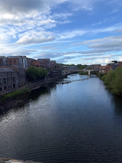 The image shows a stretch of the River Wear in Durham, United Kingdom, as viewed from an elevated position, possibly a bridge or high vantage point. The river dominates the right half of the frame, its surface dark and textured with ripples and reflections. Along the left bank of the river, a row of buildings lines the water's edge. These buildings vary in height, style, and color, ranging from older brick structures to more modern constructions with glass elements. Green foliage and trees are interspersed among the buildings, adding a touch of nature to the urban landscape. In the distance, a pedestrian bridge crosses the river. The sky above is filled with clouds, creating a dramatic backdrop with shades of blue and white.