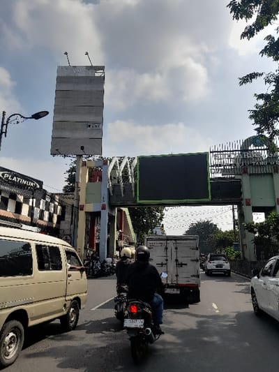 The image shows a street scene in Kecamatan Waru, Indonesia (latitude: -7.352, longitude: 112.729). A large billboard is located on the left side of the street, while a smaller billboard with a black screen is located on the right. A white van is driving on the left side of the street, and there are three motorbikes driving on the right side. The motorbikes are being driven by two males. One person is visible in the van. There are also a few cars driving in the distance. The environment is urban and the sky is partially cloudy. A street light is visible on the left side of the street.