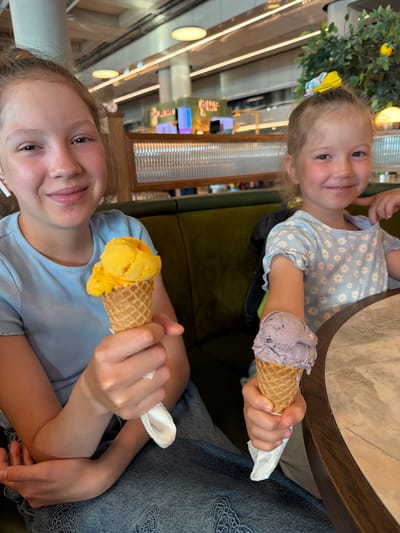 This image captures two young girls enjoying ice cream inside a modern cafe or restaurant, stated to be located in Dalaman, Turkey.

The older girl, positioned on the left, has light-colored hair, partially tied back, and is smiling broadly with visible dimples, facing the camera. She wears a light blue ribbed short-sleeved top and light blue denim jeans with a subtle, lighter-toned embroidered pattern visible on her lap. A white wireless earbud is visible in her left ear. She holds a bright yellow, two-scoop ice cream in a standard waffle cone, which is wrapped in a white napkin, in her right hand, positioned forward. Her left arm is bent, resting near her waist.

The younger girl, positioned on the right, also smiles gently at the camera. Her light-colored hair is styled in a high ponytail, secured with a bright yellow hair tie featuring a floral or patterned design. She wears a light blue short-sleeved t-shirt adorned with a pattern of small white daisies and features ruffled edges on the sleeves. She holds a two-scoop, pale lavender-colored ice cream, possibly with darker specks, in a standard waffle cone, also wrapped in a white napkin, in her left hand, positioned forward. Her right hand is partially visible, resting near her face or on the table.

Both girls are seated on dark green upholstered booth seating. A round table with a light-colored marble-patterned surface and a dark wooden rim is partially visible on the right side of the frame.

The background shows a brightly lit indoor environment with high ceilings, featuring multiple flush-mounted round and linear strip lights. White architectural pillars are visible. To the left, behind the older girl, there is a counter or partition with vertical ribbed, translucent paneling. To the right, behind the younger girl, an artificial plant or small tree with green leaves and yellow fruit, resembling lemons, serves as a decorative element. Additional blurred background elements indicate more seating areas and the general layout of a contemporary establishment.