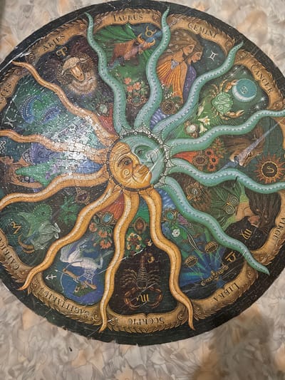 The image shows a circular jigsaw puzzle depicting the twelve signs of the zodiac. The puzzle is laid out on a textured surface that appears to be a tabletop or floor with a marble-like pattern. The central design features a sun with a face, surrounded by flowing, tentacle-like shapes in green and orange. Each zodiac sign is represented in its corresponding section of the circle, with its name written in an ornate font and a symbolic image. For example, Aries is depicted with a ram, Taurus with a bull, Gemini with two figures, and Cancer with a crab. The puzzle is intricate, with detailed artwork for each sign. The overall color palette is rich and varied, with deep blues, greens, golds, and browns. There is no indication of a specific location in the city of Айет, Kazakhstan, or any other city. The image is a close-up view of the puzzle itself, without any contextual environmental details that would place it within a specific geographical setting.