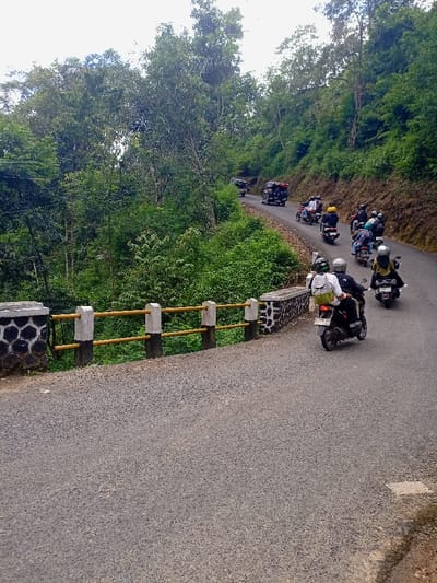 A paved road winds through a dense, verdant forest in Cisewu, Indonesia. The road descends a slope,  and a short bridge with a yellow and white railing crosses a small stream or ditch on the right side of the road.  A group of seven people are riding motorcycles up the road, and some cars are driving ahead of them in the distance. It is a cloudy day.  The coordinates of the scene are latitude -7.411, longitude 107.542.  The motorcycles are moving up the winding road, away from the bridge.  The people on the motorcycles are wearing helmets, and some are wearing jackets.  The forest is dense with green foliage, and the trees on the hillside slope down to the road.