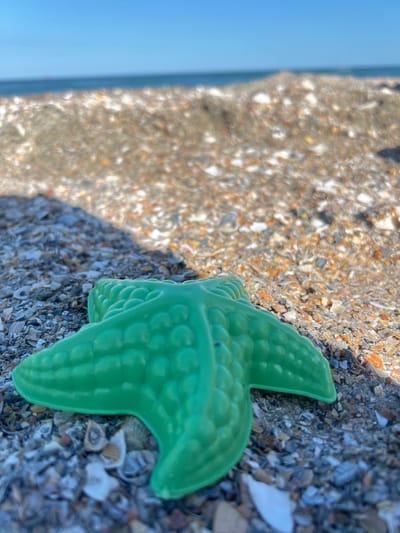 The image depicts a bright green starfish-shaped sand mold resting on a beach composed of shells and pebbles. The background shows a blurry expanse of shells, leading to a muted blue ocean and a clear blue sky. There is a distinct shadow cast on the sand mold from the left side of the frame. The scene is likely captured at a beach in Năvodari, Romania, given the context. No people or animals are visible. The lighting suggests it is daytime, possibly late morning or early afternoon.