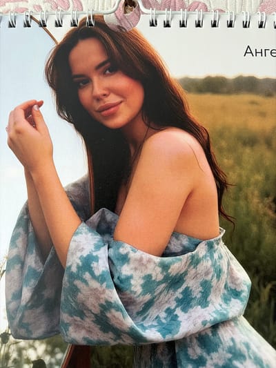 A woman with long, dark hair poses outdoors, looking towards the camera with a slight smile. She wears a blue and white patterned dress with wide sleeves. Her left arm is bent, and her hand rests near her chin. In the background, a field of tall grass and trees are visible under a bright sky. The text "Анге" is visible in the upper right corner of the image. The image is a page from a calendar, indicated by the spiral binding and perforations at the top.