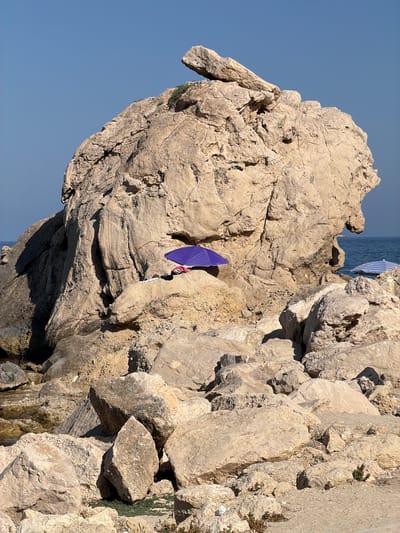 The image depicts a rocky coastline under a clear blue sky. A large, weathered rock formation dominates the center of the frame. On a ledge of this formation, a small, folded purple umbrella is open, casting a shadow. Next to the umbrella, two beach towels are visible, one white and one patterned, along with a pair of pink sandals. The rocky terrain is composed of large, light-colored boulders. In the distance, the blue ocean meets the horizon, and a sliver of another beach umbrella, striped blue and white, can be seen to the right. The scene suggests a quiet, sunny day at a secluded beach.
