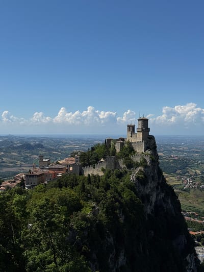 A panoramic landscape photograph captures a historic fortified city perched on a rocky mountain ridge, overlooking a vast valley. The image is presented on its side, but if rotated clockwise, it shows a clear blue sky with fluffy white cumulus clouds at the top.

In the mid-ground, distinctive medieval-style stone fortresses and towers are prominent, built upon a rugged, tree-covered outcrop. At least two large, square towers, characteristic of the Three Towers of San Marino, stand tall with crenellated walls and small flags atop. Below these fortresses, the buildings of a town or city with red-tiled roofs are nestled along the mountainside, surrounded by lush green foliage. The immediate foreground on the right (when correctly oriented) shows a steep, dark, tree-covered slope.

The background reveals an expansive valley stretching to the horizon, featuring a patchwork of cultivated fields in shades of green and brown, dotted with smaller villages and infrastructure. The bright sunlight, casting clear shadows, indicates it is daytime, likely mid-morning or afternoon, under fair weather conditions. The scene evokes a sense of history and grandeur, characteristic of the Republic of San Marino. No visible text is present in the image.