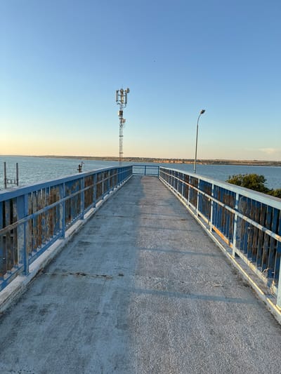 The image depicts an elevated pedestrian walkway with rusted blue railings overlooking a body of water. A cellular tower stands prominently in the distance on the left, and a lamppost is visible on the right. The walkway's concrete surface shows signs of wear. The sky is clear and blue, indicating daytime. The landscape in the background suggests a coastal or riverside setting with vegetation and distant structures, possibly within or near the city of Yelyzavetivka, Ukraine. There are no people or animals visible in the frame. The scene is static and appears to be taken from a stationary viewpoint.