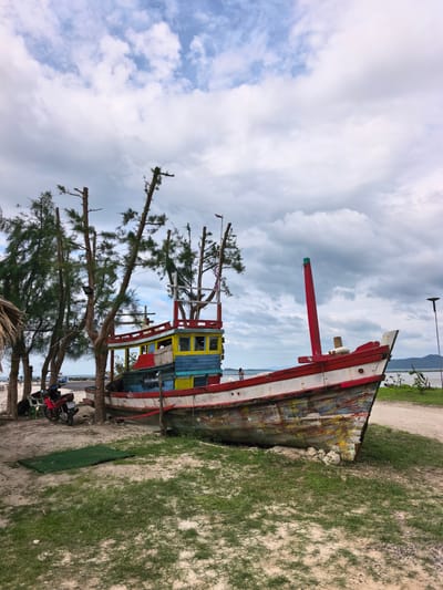 The image depicts a weathered, colorful wooden boat beached on a sandy and grassy shore in a tropical coastal setting, likely Ban Bang Khao, Thailand. The sky is overcast with large white and grey clouds, suggesting a cloudy daytime.
The primary subject is a large fishing vessel, painted in multiple vibrant, but faded and peeling, colors including red, white, blue, yellow, and green. Its bow is angled towards the right, supported by white rocks, while its stern is to the left. A tall red structure, possibly a mast or part of the hull, stands prominently near the bow. Towards the stern, there is a cabin structure painted in yellow and blue.
Around the boat, numerous tall, slender trees with sparse, needle-like foliage, possibly casuarina trees, line the shore. Many of these trees show signs of heavy pruning or damage, with several trunks cut short. The ground is a mix of light-colored sand and patches of green grass. A rectangular green mat lies on the sand to the left of the boat.
Other objects in the scene include a red motorcycle parked under a tree on the left, next to a white plastic chair. A street light pole is visible on the far right, near the water. In the distant background, across the water, low-lying hills or mountains are visible under the cloudy sky.
Two people are present in the scene. One is faintly visible on the boat's deck near the stern, and another small figure stands by the water's edge to the right of the boat. No specific activity is clearly depicted; the boat appears to be stationary, possibly out of use or undergoing repair.
A license plate on the red motorcycle is partially visible, showing "2083" and "4115" along with Thai script, which likely indicates its registration.