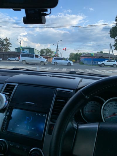 The image is taken from inside a vehicle, looking forward through the windshield. A dashboard, steering wheel, and instrument cluster are visible in the foreground. Outside the vehicle, a street intersection with traffic lights and crosswalks is depicted. Several vehicles are present, including a silver minivan, a white crossover SUV, and another silver minivan. Pedestrian crossing signs are visible on poles. Buildings and trees line the street, and the sky is cloudy. A dashcam is mounted on the windshield. The scene is consistent with a traffic situation in a city, possibly Belogorsk, Russia, given the information.