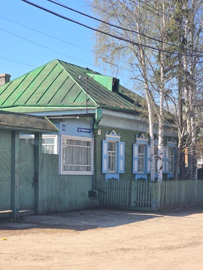 The image captures a side view of a weathered, two-story wooden house in Lensk, Russia. The house is painted green, with a green metal roof, and features decorative blue window shutters. A street sign, reading "ул. Аммосова," and the number "22" are affixed to the exterior. Birch trees with bare branches stand near the house, partially obscuring the view of the upper floors. Electrical wires run overhead. To the right of the house is a dirt road or pathway, cast in shadow. The sky is clear and blue.