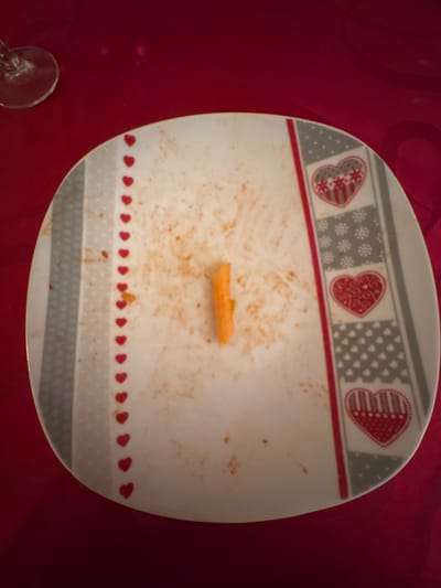 A nearly empty white plate sits on a red surface. The plate's edges are decorated with hearts and geometric patterns. Remnants of food stains and a single piece of pasta remain. A glass stem, possibly part of a wine glass, is visible at the lower left corner of the image. The plate appears to be a decorative piece and has a distinctive border design.