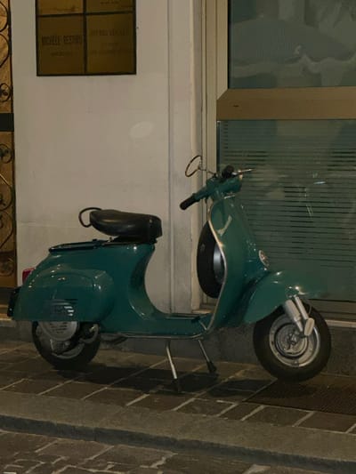 A teal-green vintage Vespa scooter is parked on a brick-patterned paved sidewalk in front of a building facade in the city of Pavia, Italy. The scooter faces right, supported by its kickstand. It features a black single-rider seat with a small grab handle behind it, chrome handlebars with circular mirrors on each side (only the right is clearly visible), and a spare tire mounted centrally on the front cowl. The front wheel and suspension components are visible. The building's white-painted wall is visible behind the scooter. To the left, a portion of an ornate golden metal gate or door is present. Above the scooter's rear, a rectangular bronze-colored plaque is mounted on the wall. The plaque is divided into quadrants, with legible text in the upper left reading "MICHELE RESTIVO" and below it "AVVOCATO". The upper right quadrant reads "STUDIO LEGALE". To the right of the scooter, a large window or glass door with a horizontal metal frame and vertical blinds is visible. The scene is illuminated, suggesting nighttime or indoor conditions. No individuals are present.