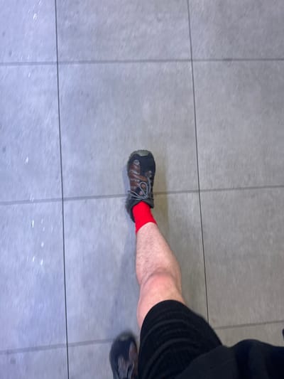 A high-angle, slightly blurry photograph shows a person standing on a tiled surface. The person is wearing black shorts, red socks, and dark hiking shoes. The surface appears to be light grey tiles with dark grout lines. The scene suggests an indoor setting, possibly a public space. There is no information to determine its location in Uttlesford, United Kingdom.