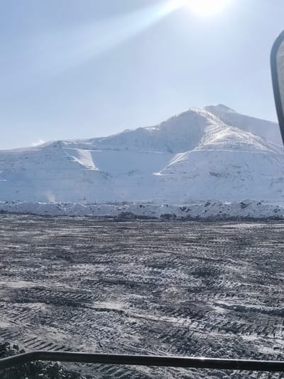 The image presents a vast open-pit mining or industrial scene situated in a snow-covered mountainous region near Im. Gastello, Russia, under a bright, clear sky. The upper third of the frame shows a light blue sky with a prominent solar glare emanating from the upper right. A large, snow-covered mountain range occupies the mid-ground, characterized by distinct artificial terraces or benches indicative of excavation. Sparse, dark coniferous trees are visible on some snow-clad slopes. In the distance, beyond a long, irregular snow berm that traverses the mid-ground, faint industrial structures are discernible. The foreground consists of an expansive, flat, dark gray to black ground surface, partially covered with patches of snow or frost. This surface is heavily marked with numerous deep tire tracks from heavy vehicles, suggesting active operations. Small piles of dark, granular material are scattered on the foreground. A dark, horizontal metallic bar or railing is present at the bottom edge of the frame, indicating an elevated perspective. No individuals are visible. The scene conveys cold, winter conditions with ongoing large-scale industrial activity.