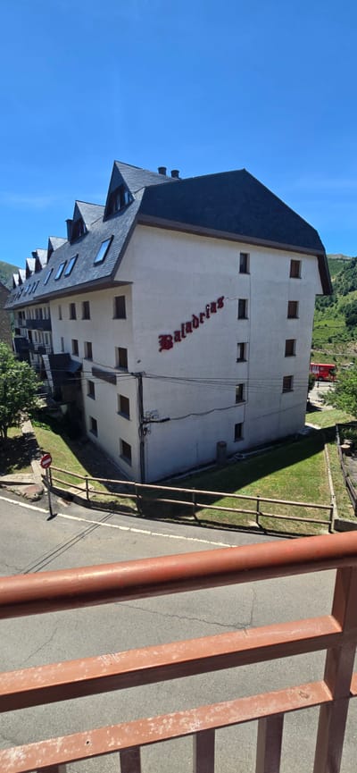This image depicts a multi-story building with a dark sloped roof, characteristic of mountain architecture. The building's exterior walls are a light color, and it has several rectangular windows. Prominently displayed on one side of the building is the name "Baladrigs" in red, stylized lettering. The building appears to be situated in a hilly or mountainous area, as suggested by the green slopes and trees visible in the background. A red vehicle is partially visible behind the building. A metal railing, painted in a reddish-brown hue, runs across the foreground, partially obscuring a paved road or pathway. The sky is a clear, bright blue, indicating it is daytime with sunny weather. The scene appears to be a quiet, possibly rural, location in Sallent de Gállego, Spain. There are no people visible in the image.