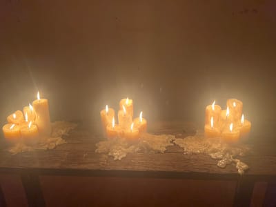 Three groups of lit, thick, yellow candles stand on a weathered wooden surface. Each cluster spills abundant wax, solidified in irregular shapes beneath. The background is a plain, shadowed wall. The lighting is low, emphasizing the candle flames and wax textures. There's a sense of warmth and possible antiquity to the scene.