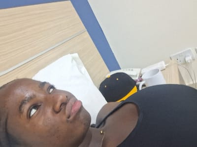 A single individual with dark skin is depicted lying supine on a bed, their head resting on a white pillow. The individual is wearing a dark, sleeveless top and a delicate gold-toned necklace. Their gaze is directed upwards and to the left, with lips slightly pursed. The background features a light wood-paneled headboard with a blue accent strip above it. To the individual's right, on a bedside surface, a black and yellow baseball cap, a white ceramic mug, a white corded telephone, and a white tube dispenser are present. An electrical wall outlet with a white plug and attached wires is visible on the adjacent wall. The scene is an indoor setting, likely a bedroom or hotel room, located in Close, Nigeria.
