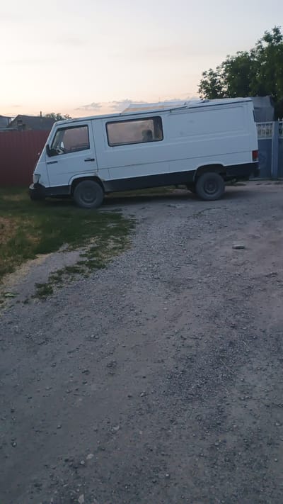 The image shows a white panel van parked on a gravel road next to a grassy area. The van is the primary object and occupies the left side of the frame. The scene is outdoors, with a clear sky visible in the background, suggesting it is daytime, likely late afternoon or early evening given the soft light. A few trees are also visible in the distance, and what appears to be a building with windows is to the left of the van.
The van is white with black trim around the windows and doors. It has two doors visible at the front and appears to be a long-wheelbase model. The wheels of the van are black. The gravel road surface is textured with small stones and dirt. The grassy area next to the road shows some green grass, interspersed with some dry or brown patches.
There is no discernible activity or event taking place; the van is parked. Notable details include the lighting, which is diffused and soft, typical of a sunset or sunrise. The overall mood conveyed by the image is peaceful and quiet. There is no visible text in the image. The location context provided is Mitoc, Moldova, but there are no specific visual cues in the image to confirm this location.