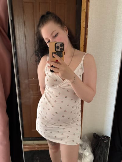 A young woman with dark hair pulled into a ponytail is taking a selfie in a mirror. She is wearing a white slip dress with a floral print. She is holding a smartphone in her right hand, and a ring is visible on her right ring finger. A silver bracelet is on her left wrist. The background appears to be an interior setting with a wooden door and a textured wall. A large mirror is positioned to her left, reflecting her. To her right, there are two trash bags. The image appears to have been taken in the city of Vylgort, Russia.