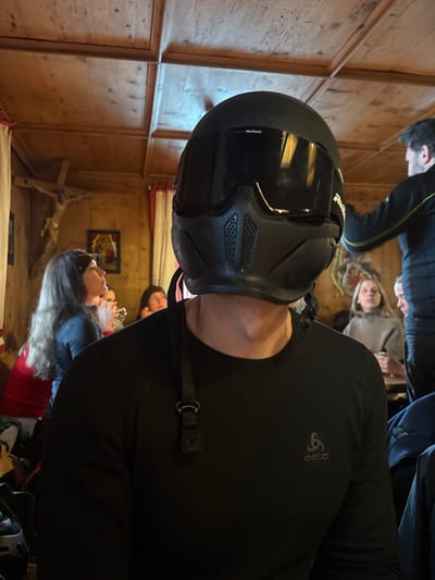 An individual wearing a matte black full-face helmet with a dark visor and integrated chin bar ventilation is positioned centrally, facing forward in an indoor setting. A black strap from the helmet hangs over their right shoulder. The person wears a black Odlo long-sleeve athletic shirt.
The background reveals a rustic, wood-paneled interior, characteristic of an alpine lodge or restaurant in St. Christoph, Austria. The walls are constructed of light-toned wood, and the ceiling features a coffered wood design.
On the left side of the background, a large wooden crucifix is affixed to the wall. Below it, a framed artwork is visible. A woman with long, dark hair, wearing a dark top, is seated in the mid-ground, looking to her right, seemingly engaged in conversation. Red fabric, possibly a curtain or drapery, is present near the wall.
On the right side of the background, a man with dark hair, wearing a dark top with light-colored trim, stands with his back partially to the camera, appearing to be in motion or reaching for something. Further right, a woman with light hair, wearing a beige turtleneck top, is seated at a table, looking to her right. Other indistinct figures and partial views of additional furnishings are visible in the mid-to-far background. The overall scene suggests a social gathering within a traditional wooden establishment.