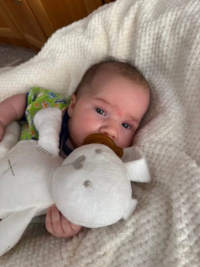 A newborn baby with fair skin and light brown hair is lying on a white, textured blanket. The baby is wearing a green onesie with a car print and a blue collar. A stuffed toy with grey embroidered features is next to the baby, and the baby's pacifier is in its mouth. The baby's left hand is visible, resting on the stuffed toy. The background is blurred, showing a wooden surface and a textured surface. This image was likely taken indoors. There is no discernible information about the location in the city of Выльгорт, Russia.