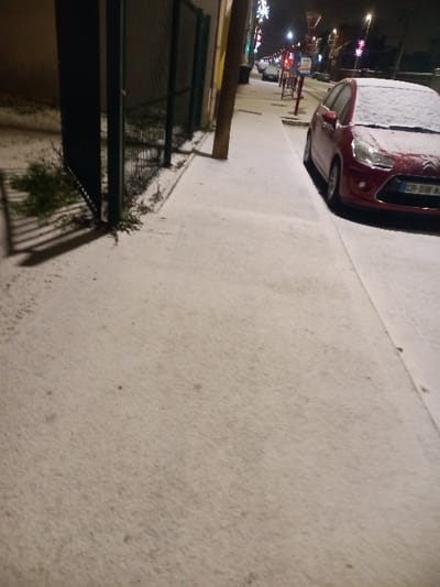 A snowy street scene at night. A burgundy car parked partially on the snowy sidewalk, covered in snow. A green fence and vegetation are on the left side. Distant blurred lights suggest a town or city street. The ground and surfaces are covered in a layer of snow, indicating a recent snowfall.