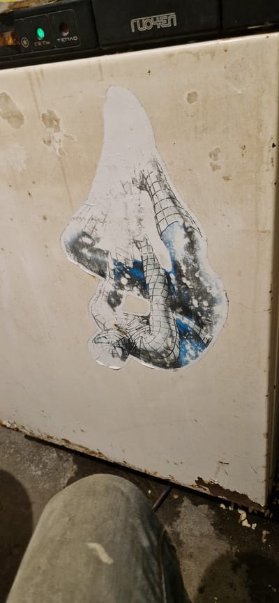 A partially peeled sticker of the Marvel character Spider-Man is affixed to the side of a white appliance, possibly a refrigerator. The sticker depicts Spider-Man in a crouched or swinging pose, with his web-patterned suit visible. The background of the appliance is a beige or off-white color and shows signs of wear, with some discoloration and rust spots, particularly along the bottom edge. The appliance appears to be located in an indoor environment, possibly a basement or utility room, given the concrete floor and visible pipes or wires in the lower portion of the image. A person's legs, clad in blue jeans, are visible in the foreground, suggesting they are standing or sitting near the appliance. The top of the appliance has a black panel with some labels and indicator lights. The word "ГУОЧЕН" is visible on this black panel. The overall scene suggests a domestic or slightly neglected setting.