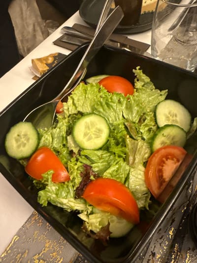 A black dish holds a fresh salad. Green lettuce forms the base, adorned with sliced cucumbers and ripe tomatoes. The cucumbers display a light green hue and visible seeds. The tomatoes are red and partially sliced. Silver serving utensils rest within the dish, suggesting the salad is ready to be served. The dish sits on a surface with decorative elements.