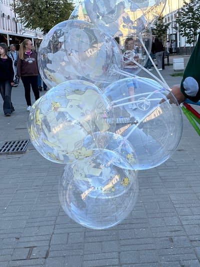 The image depicts a cluster of large, transparent inflatable globes, some adorned with images of Pikachu. They are held aloft by unseen strings attached to a set of wands, one of which is partially visible in the lower right corner being held by a person. In the background, several individuals are walking on a paved walkway. The setting appears to be an outdoor urban environment, possibly a street or plaza, with buildings and trees visible in the distance. The ground is paved with interlocking grey bricks. This scene is located in the city of Feklino, Russia.
