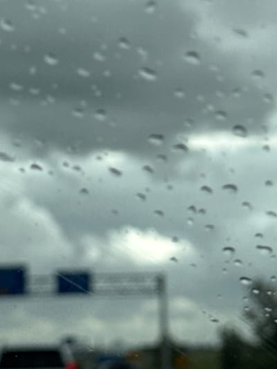 The image depicts a rain-streaked windshield with a blurred view of an overcast sky and some objects in the distance. In the background, two dark rectangular signs are visible, likely traffic or informational signs, mounted on a pole. A portion of a vehicle's hood is also discernible at the bottom. The scene appears to be from a roadside or highway setting, suggesting a journey in inclement weather. The location is Marsum, Netherlands.