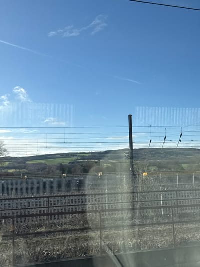 Captured through a window, a bright blue sky contrasts with the landscape beyond. A prominent contrail streaks across the sky. Parallel railway lines dominate the right side of the frame, running adjacent to a green, undulating terrain. Overhead power lines stretch across the scene, supported by dark poles. The image suggests a view from a moving train.
