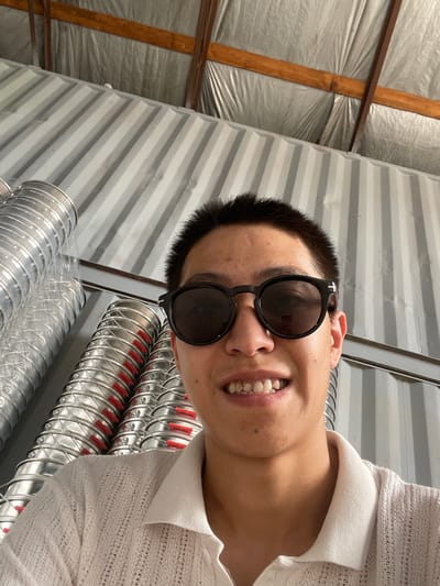 The image shows a close-up, low-angle selfie of a young man wearing sunglasses and a white collared shirt. He is smiling, revealing his teeth. The background consists of industrial elements, including corrugated metal walls and large, coiled silver ventilation ducts with red accents. The ceiling above appears to be made of wood beams and reflective insulation. The scene suggests an indoor, possibly industrial or storage setting within Кенен Әзірбаев ауылы, Kazakhstan.