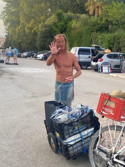 The image depicts an outdoor scene at Playa Del Sol-Villacana, Spain, likely a parking area near the beach. A shirtless man with long blonde hair, wearing a blue sarong and holding a cigarette, stands in the foreground facing the camera and waving with his left hand. He appears to be speaking. To his left, there is a black cart with two stacked crates, one filled with a blue and white patterned blanket and other items, and the other containing a mattress. To the right of the cart, a red Coca-Cola crate is visible, mounted on the back of a bicycle. In the background, several cars are parked, and lush green trees and foliage form a backdrop. Some people are seen in the distance near the cars. The ground is paved with asphalt.