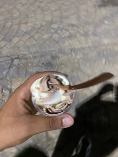 A person's hand holds a clear plastic cup filled with ice cream. The ice cream is swirled with at least two colors, appearing to be chocolate and vanilla or a light cream color. A wooden spoon is partially submerged in the ice cream, with its handle extending out of the cup and angled to the right. The background is a grey, textured surface that appears to be pavement, possibly cobblestone or interlocking pavers, with a faint pattern. There is a dark shadow cast on the lower right side of the image. The image appears to be taken outdoors in an urban or paved area. The location is identified as Tipaza, Algeria.