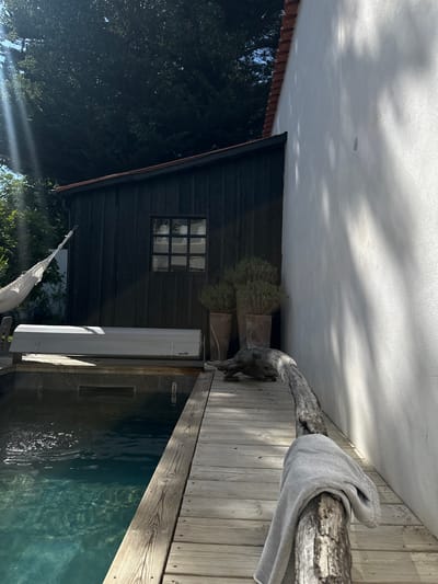 The image depicts an outdoor scene with a building facade, a wooden deck, and a body of water, possibly a pool, adjacent to the deck. The building has a dark, likely wooden, exterior with a multi-paned window and two potted plants near the roofline. A white, rectangular object, perhaps an air conditioning unit or storage container, is situated close to the building.
The deck is constructed of light-colored wooden planks. A gnarled tree branch serves as a decorative element or a railing along the edge of the deck, with a gray garment draped over it.
The pool has a dark, slightly reflective surface. The sun is visible in the lower left corner of the image, suggesting it is daytime.
While the precise location cannot be confirmed from the image alone, the prompt indicates La Couarde-sur-Mer, France as the likely setting.