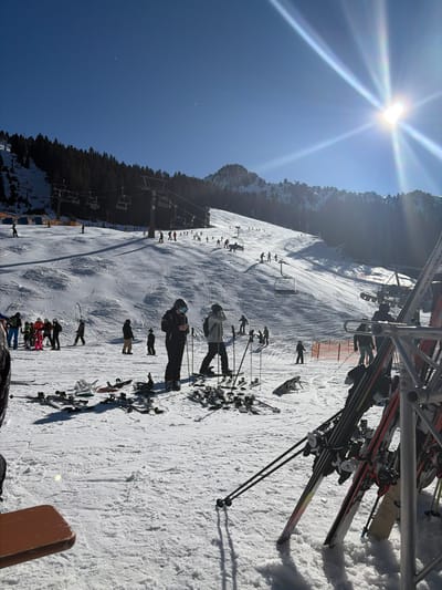 The scene depicts a busy ski resort on a bright, sunny day in Schwendau, Austria. The foreground features a snow-covered ground with numerous pairs of skis and several snowboards laid flat. Multiple ski poles are stuck upright in the snow. To the right, a metal ski rack holds several pairs of skis vertically. A brown wooden table edge is partially visible in the bottom-left corner.

In the mid-ground, several individuals are present. One person in dark ski attire, wearing a helmet and goggles, stands holding ski poles, appearing to look at a smartphone. Another individual in a light-colored jacket and dark pants stands on skis, also wearing a helmet and goggles, with ski poles. Other people in winter gear are scattered across this area, some preparing or resting.

The background reveals a wide, snow-covered ski slope with numerous skiers and snowboarders actively descending or standing. Several ski lift towers and operational chairlifts are visible across the slope. A dense pine forest covers the upper left slope, and snowy mountain peaks are discernible in the far distance. A bright sun, exhibiting prominent lens flares, is positioned in the upper-right clear blue sky. An orange safety net is partially visible on the right side of the main slope.