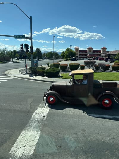 A person drives a rusty, vintage pickup truck with red rims down a street in Fernley, United States of America. The truck is a dark brown color and appears to be missing some of its original body panels. The driver, wearing sunglasses and a white shirt, is smiling and giving a thumbs up. The truck is in the foreground of the image, crossing a cracked white crosswalk line. In the background, a Super 8 hotel with a distinctive red roof is visible, along with a Chevron gas station sign. Traffic lights and street signs, including one indicating "US 95A," are also present. The sky is blue with scattered white clouds, and the sun is shining. The environment appears to be an urban or suburban setting with some green landscaping and trees.