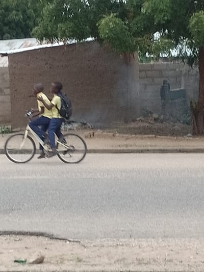 Two young individuals, dressed in yellow shirts and blue pants, are riding a white bicycle along an asphalt road. The rider in front is pedaling while the passenger in the back is holding onto the rider. The passenger is wearing a black backpack. The setting appears to be in a residential area of Biliri, Nigeria. In the background, there's a rudimentary building with a corrugated metal roof, a wall, a tree, and a partially obscured structure. A green bottle is present on the ground.