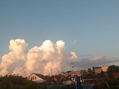 In the city of Verkhnevesyoloye, Russia, a sunset illuminates the sky with warm hues casting a golden glow on a cluster of cumulus clouds. The scene features a residential area nestled on a hillside, with houses of varying architectural styles and sizes visible. Amidst the buildings, two red and white striped power transmission towers stand prominently, their structures reaching towards the sky. Lush green trees and foliage surround the houses, adding a natural element to the urban landscape. In the foreground, a metal fence encloses the area, with a glimpse of a blue industrial container with lettering visible. The overall atmosphere is serene, capturing a moment of natural beauty over a populated area.