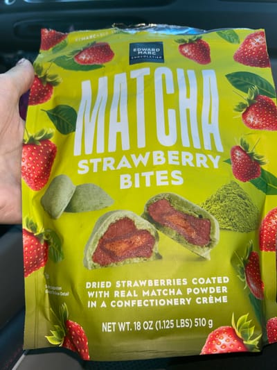 A person's hand is holding a bag of Edward Marc Chocolatier Matcha Strawberry Bites in Everett, Washington, USA. The bag is bright yellow and features images of strawberries, green leaves, and matcha powder. Text on the bag indicates "MATCHA STRAWBERRY BITES" and "DRIED STRAWBERRIES COATED WITH REAL MATCHA POWDER IN A CONFECTIONERY CREME". The weight is listed as "NET WT. 18 OZ (1.125 LBS) 510 g". The background appears to be the interior of a car.