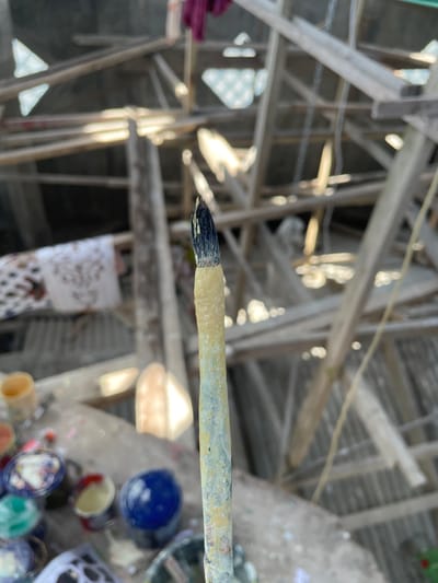 The image captures a close-up of a well-used paintbrush, held upright and centered in the foreground, against a blurred backdrop of an outdoor or semi-outdoor workshop in Kumbang, Indonesia. The paintbrush has dark bristles showing traces of yellow paint, and its handle is thickly coated with dried, light yellow/tan paint, with visible streaks of other colors. In the lower-left foreground, a collection of small, open containers, including ceramic vessels and plastic pots, hold various colored paints or glazes, such as blue, red, yellow, and green. The background reveals a complex, indistinct structure composed of crisscrossing light-colored wooden beams and scaffolding, consistent with an unfinished building or a craft area. Patches of bright sunlight illuminate parts of this structure. Hanging within the background, blurred, are pieces of fabric, including a dark red cloth and a white patterned cloth. No individuals are visible in the scene.