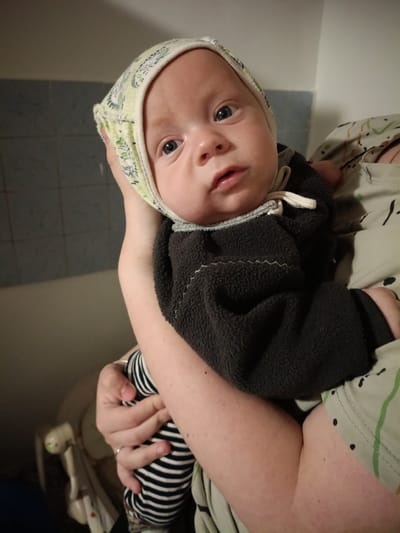A close-up, medium shot captures a baby being held by a person. The baby, wearing a patterned bonnet and a dark fleece garment over striped leggings, is positioned in the center of the frame. The baby's face is visible, with wide blue eyes looking forward, and their lips are slightly parted. The person holding the baby is partially visible, with their arms supporting the infant and their patterned clothing evident. The background consists of a tiled wall and a lighter colored wall, suggesting an indoor environment. The scene's lighting is warm and slightly dim. The location in the city of Litynia, Ukraine, is not discernible from the visual information provided in the image.