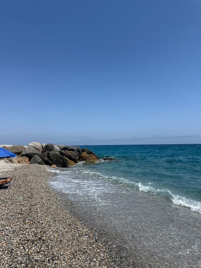 A wide-angle, eye-level shot captures a tranquil scene at a pebble beach in Finale Ligure, Italy. The clear blue sky dominates the upper portion of the frame, with a faint haze on the horizon where it meets the equally clear turquoise ocean. A breakwater, constructed of large, rough-hewn rocks, juts out into the water from the left side of the image, its base met by gentle waves. The beach itself is covered in small, smooth pebbles of varying shades of gray, brown, and white.

In the foreground, to the left, a glimpse of a person's legs and feet are visible, stretched out on a lounge chair partially obscured by a bright blue beach umbrella. The umbrella casts a shadow on the pebble shore. The water's edge is defined by small, foamy waves washing over the pebbles, creating a shallow, transparent layer. In the distance, a small sailboat can be seen on the vast expanse of the sea. The overall impression is one of peace and relaxation on a sunny day.