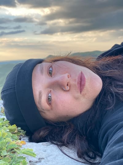 A close-up, upside-down, head-and-shoulders shot of a person lying on their back on rocks outdoors. The person has light skin, freckles, light brown hair, and green eyes, wearing a black beanie and a black hooded jacket. They have a silver hoop piercing in their left nostril and are looking upwards. In the foreground, there are green plants and light-colored rocks. In the background, there are blurred hills and a cloudy sky. The scene appears to be located in a natural, mountainous area, possibly near Абзаново, Russia.