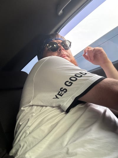 The image shows a close-up of a person's torso and arm, with their head slightly visible. The person is wearing a white t-shirt with black trim and black text that reads "YES GOOD". They are also wearing sunglasses and a blue bandana around their head. The person's skin is fair, and they have reddish-brown hair and facial hair.

The setting appears to be inside a vehicle, possibly a car, with a sunroof open above. The sky is visible through the sunroof, indicating it is daytime. The lighting suggests it might be a sunny day, but the image is somewhat underexposed, making it difficult to determine the exact time of day or weather conditions. The person seems relaxed, with their arm resting on their leg. The focus is on the t-shirt and the person's upper body, with the background blurred. The image is shot from a low angle, looking upwards.