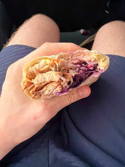 A close-up shot shows a person's hand holding a half-eaten chicken wrap. The wrap is made of a light-colored tortilla and filled with shredded chicken, coleslaw, and red cabbage. The background is blurred, but appears to be indoors, with a dark surface on the left and a person's legs and blue fabric on the right. The lighting suggests it is daytime. The overall scene is casual and appears to be a person eating on the go. There is no visible text in the image.