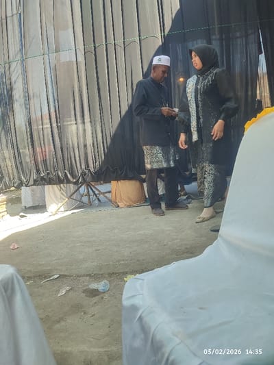 Two people stand in front of a black curtain backdrop. The person on the left wears a white Muslim prayer cap, a black jacket over a traditional patterned garment, and dark pants. The person on the right wears a black hijab and a matching black and patterned outfit. Both appear to be engaged in a conversation. The foreground is dominated by white covered chairs, suggesting an event setup. Debris litters the ground.