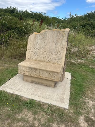 This is a close-up, eye-level, outdoor, vertical shot of a carved stone sculpture. The sculpture is light-colored, appearing to be made of sandstone or a similar material. It is set on a concrete base in a grassy, somewhat overgrown area. The background consists of lush green foliage and a bright blue sky with scattered white clouds, suggesting it is daytime and likely a fair-weather day.
The sculpture itself is intricately carved with various floral motifs, including daisies and other flowers, as well as depictions of insects like butterflies and beetles. On the side of the sculpture, there are also carvings of what appear to be animals or mythical creatures, one resembling a snake or dragon, and another with spiral designs. The carvings are shallow but detailed, giving the stone a textured appearance. The sculpture is weathered, with some areas showing moss or lichen growth.
The scene is tranquil and appears to be in a natural or semi-natural setting, possibly a park, garden, or countryside location. The Borough of Swale context suggests this could be a public art installation or a historical marker within that region of the United Kingdom. No people are visible in the image. The time of day is indicated by the bright sky and shadows, suggesting it is likely mid-morning or afternoon. There is no visible text on the sculpture or in the surrounding environment. The overall impression is one of a charming and artistic detail within a natural landscape.