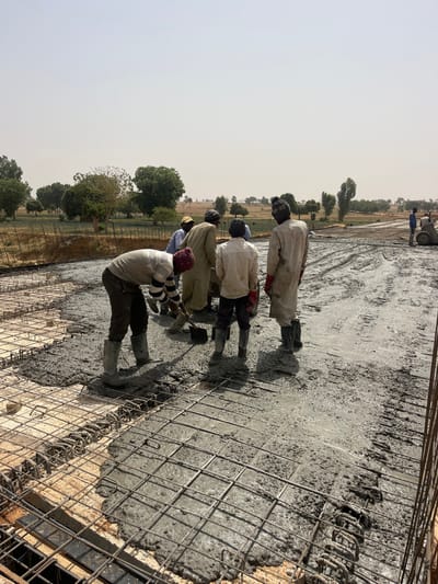 This image captures an outdoor concrete pouring operation in Gyellesu, Nigeria, during daylight hours under a hazy sky. In the foreground and center, a construction crew consisting of approximately six to seven adult males is actively working on a large, flat concrete slab.
The primary subject in the immediate foreground is a male worker bent over, wearing a light-colored striped long-sleeved shirt, dark trousers, a maroon beanie, and grey rubber boots. He is actively using a shovel to spread wet, grey concrete over an exposed grid of steel rebar. Sections of the rebar grid are visible both under the fresh concrete and in adjacent areas where the pour is progressing, revealing the underlying formwork.
To the right and slightly behind this worker, a group of four other men stands in close proximity. One individual wears a light blue shirt and a yellow cap. Another wears light-colored traditional attire and a turban. A third, facing away from the viewer, wears a light-colored, long-sleeved garment, dark boots, and red gloves. Further back on the right, another worker is visible near a piece of wheeled construction equipment, possibly a concrete mixer or a large wheelbarrow, situated on a concrete-splattered, muddy surface.
The background consists of a rural or semi-rural landscape featuring flat, dry fields bordered by scattered green trees, extending towards a distant, flat horizon under the bright, hazy sky. The scene depicts a manual construction effort focused on the creation of a reinforced concrete structure.